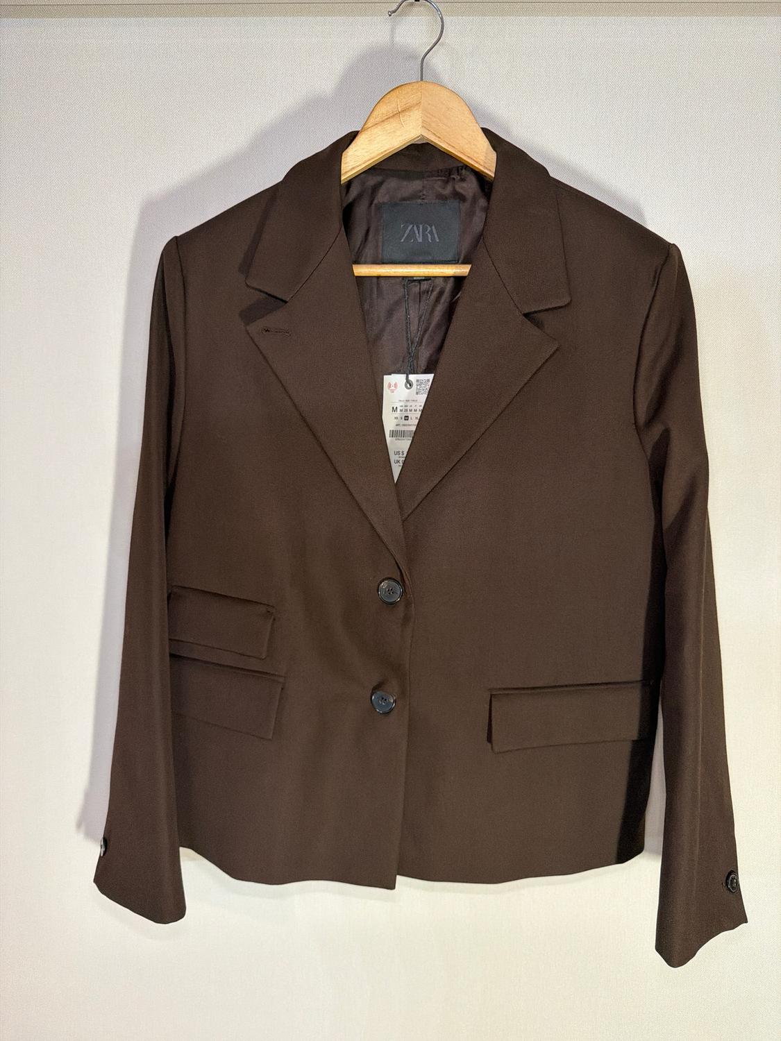 Two-button brown jacket hanging on a wooden hanger, with a visible label inside and a white background.