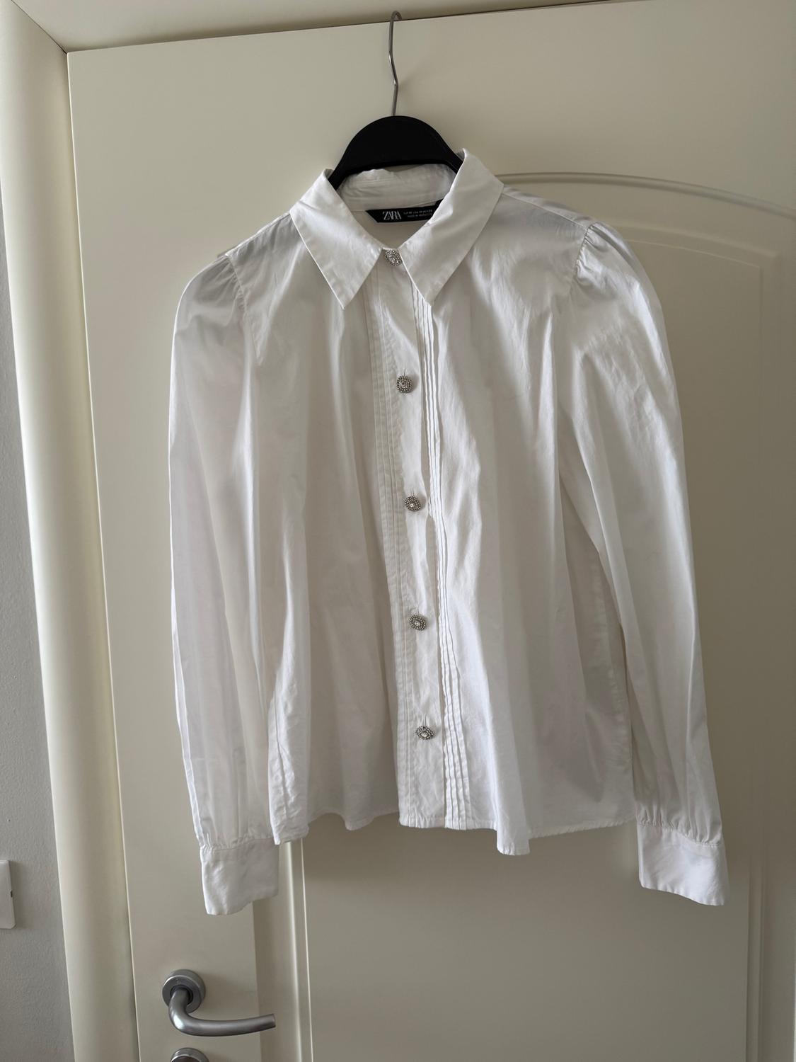 White long-sleeved shirt hanging on a black hanger over a white door. Decorative buttons visible on the front.