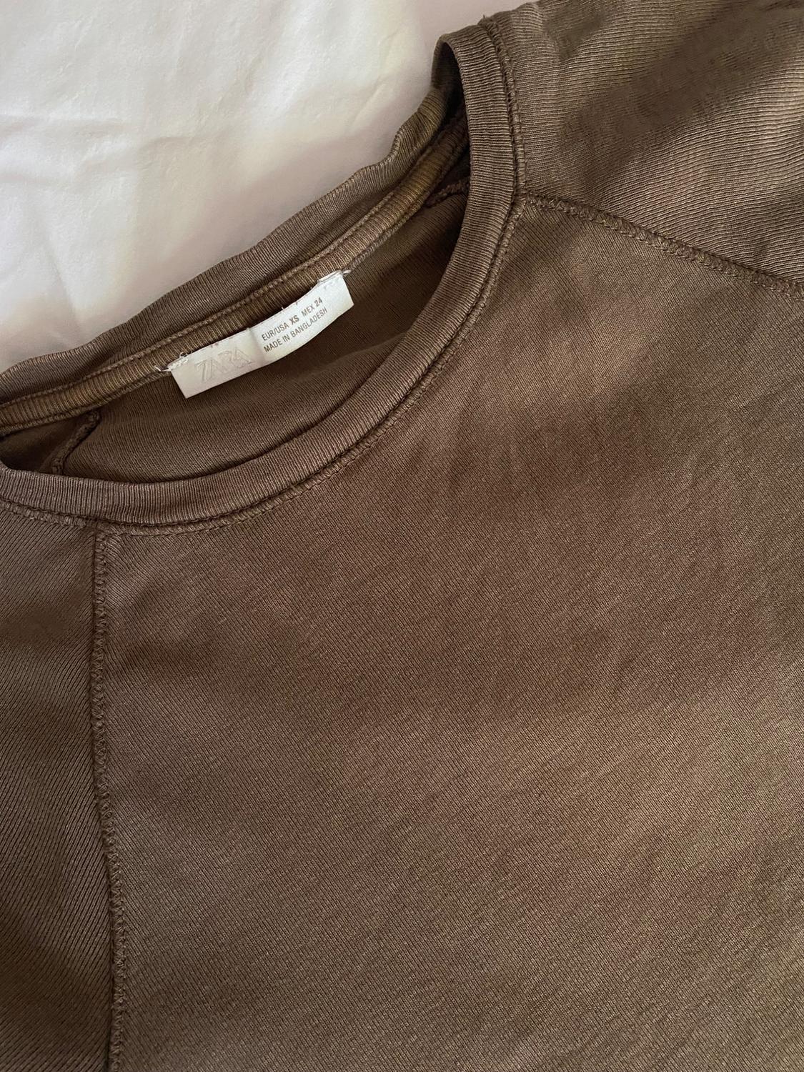 Brown round-neck t-shirt detail on a white background. Label: ZARA EUR/USA XS MEX 24 MADE IN BANGLADESH.