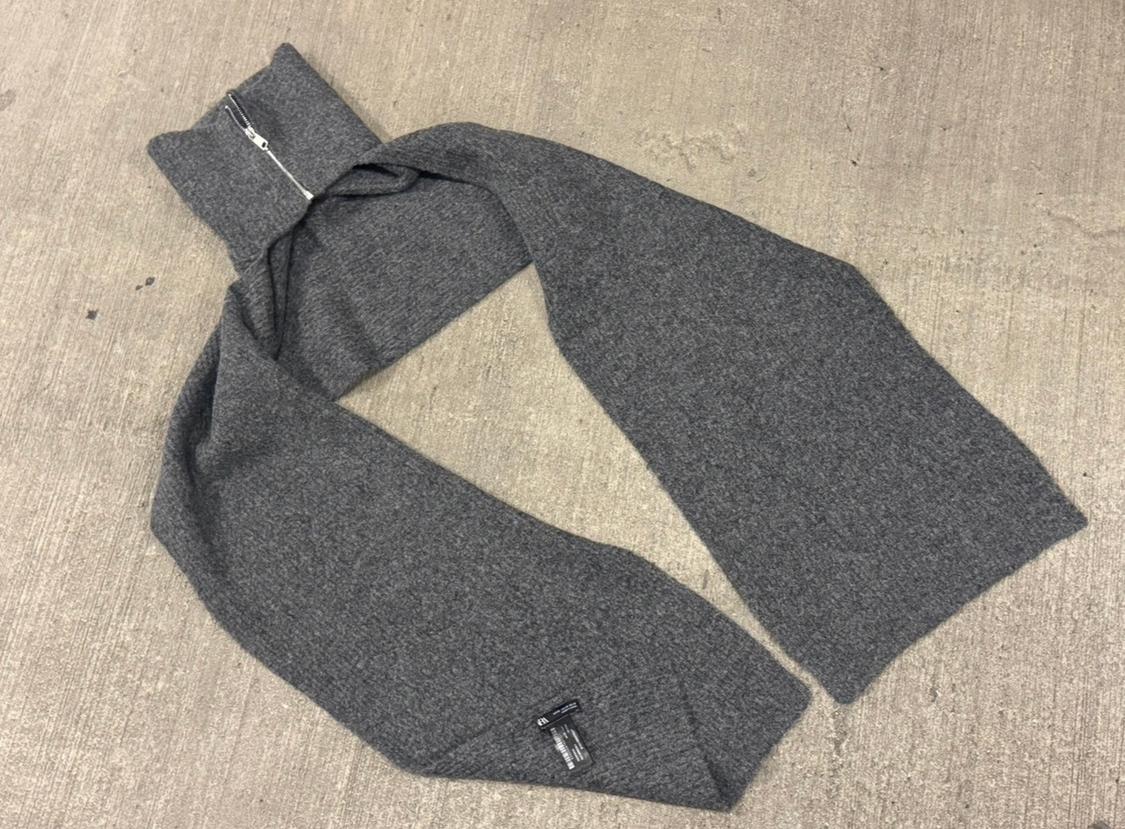 Gray wool scarf with a zipper at one end, spread out on a rough-textured floor. Label visible on the edge.