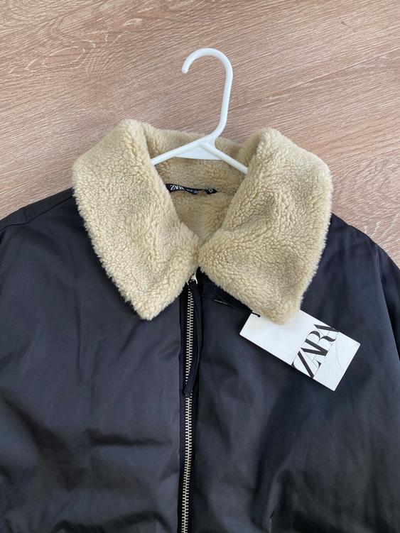 Black jacket with zipper and beige shearling collar, hanging on a white hanger. Visible label with text "ZARA".