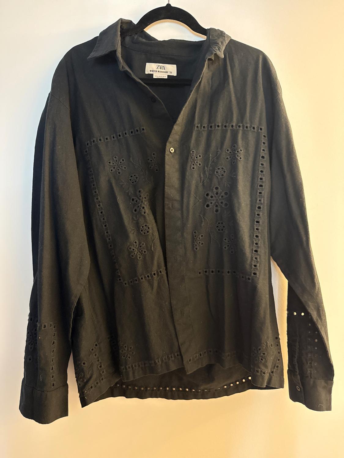 Black shirt with floral embroidery and perforations, hanging on a hanger. Visible tag: "ZARA Winter Wardrobe '23".