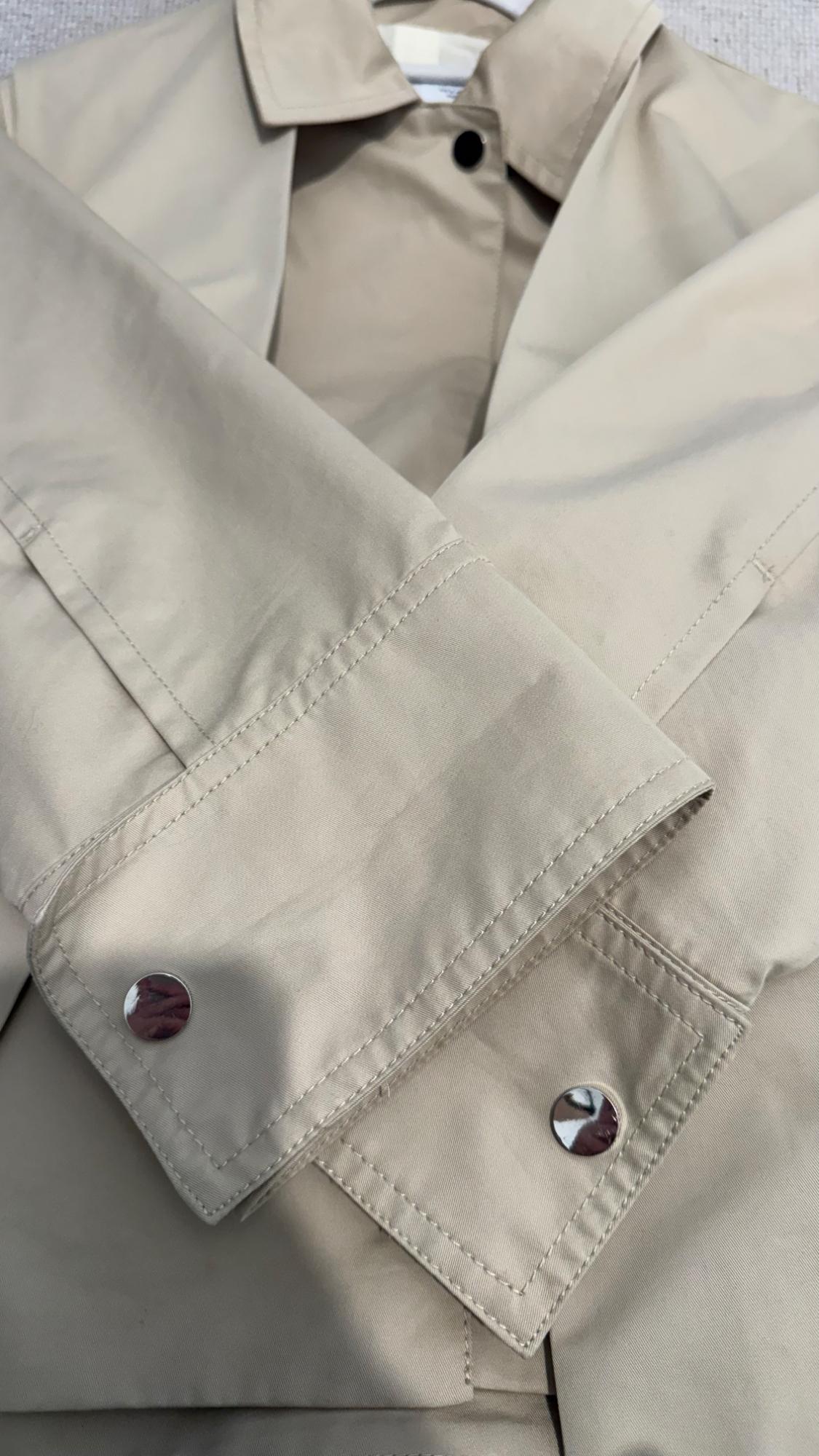Detail of a beige coat with lapels and metallic buttons. The fabric has a smooth finish and visible seams.