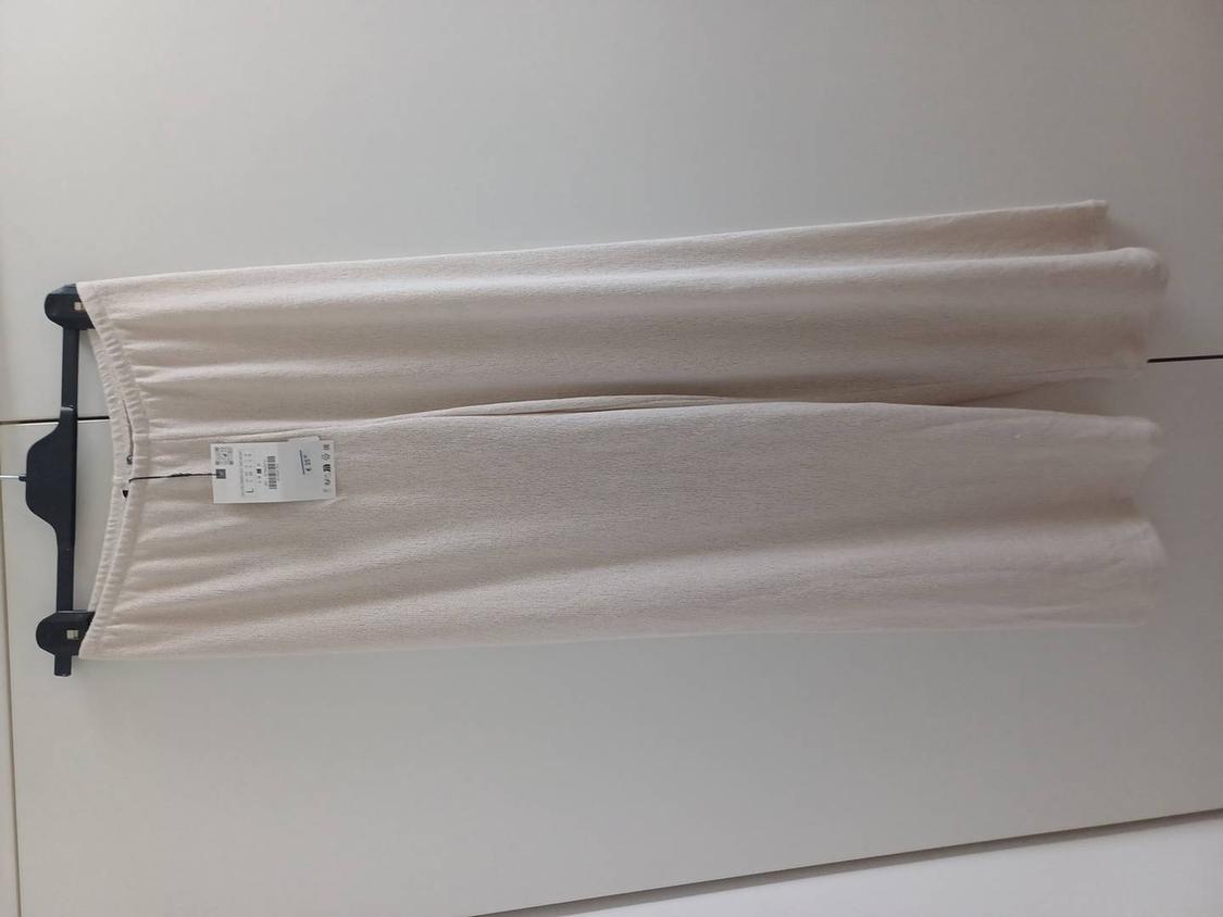 Beige trousers hanging on a black hanger with a visible tag. Plain white background.