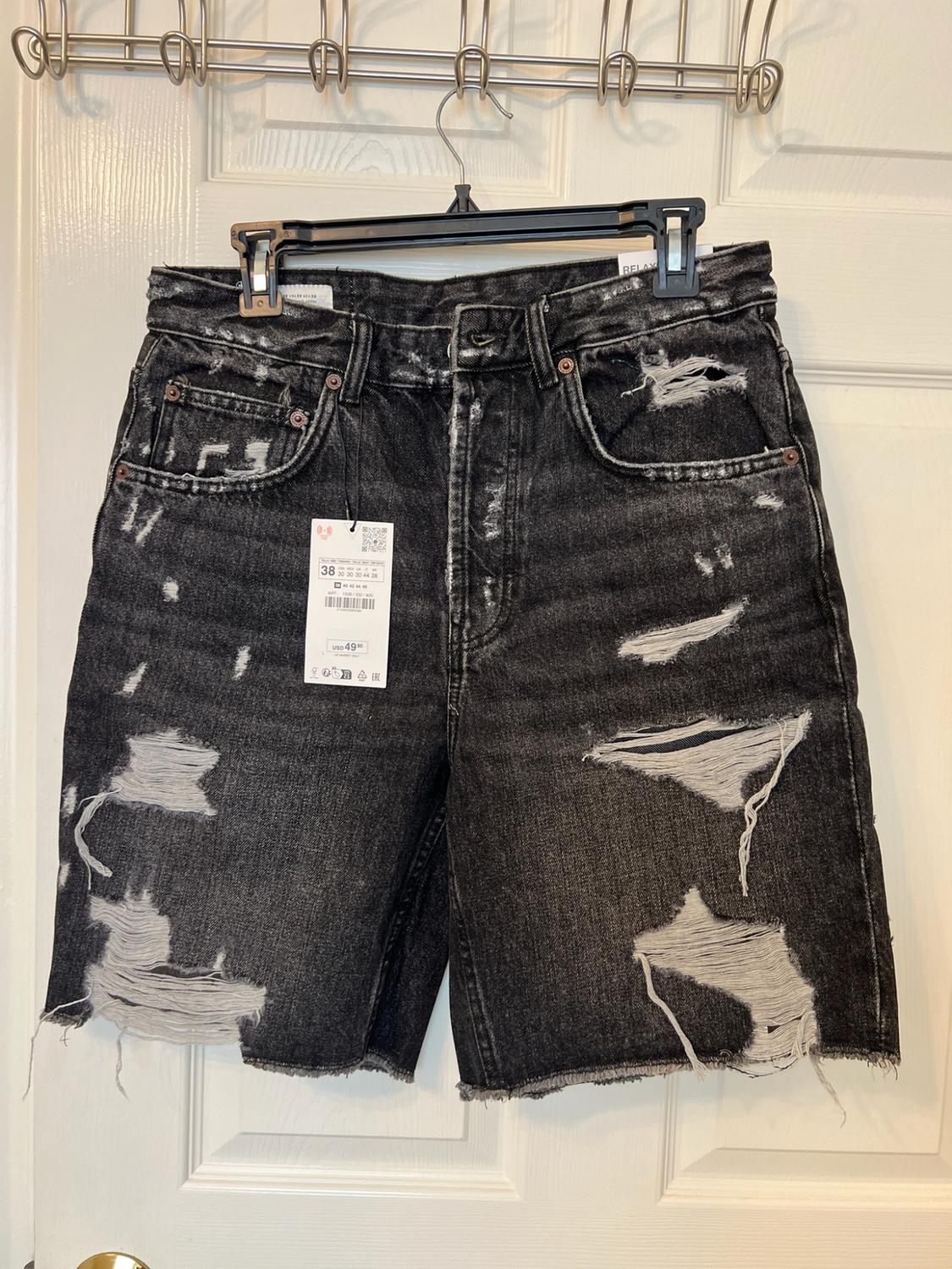 Distressed black denim shorts with rips hanging on a white door. Visible text: 38, USD 49.90, RELAX.