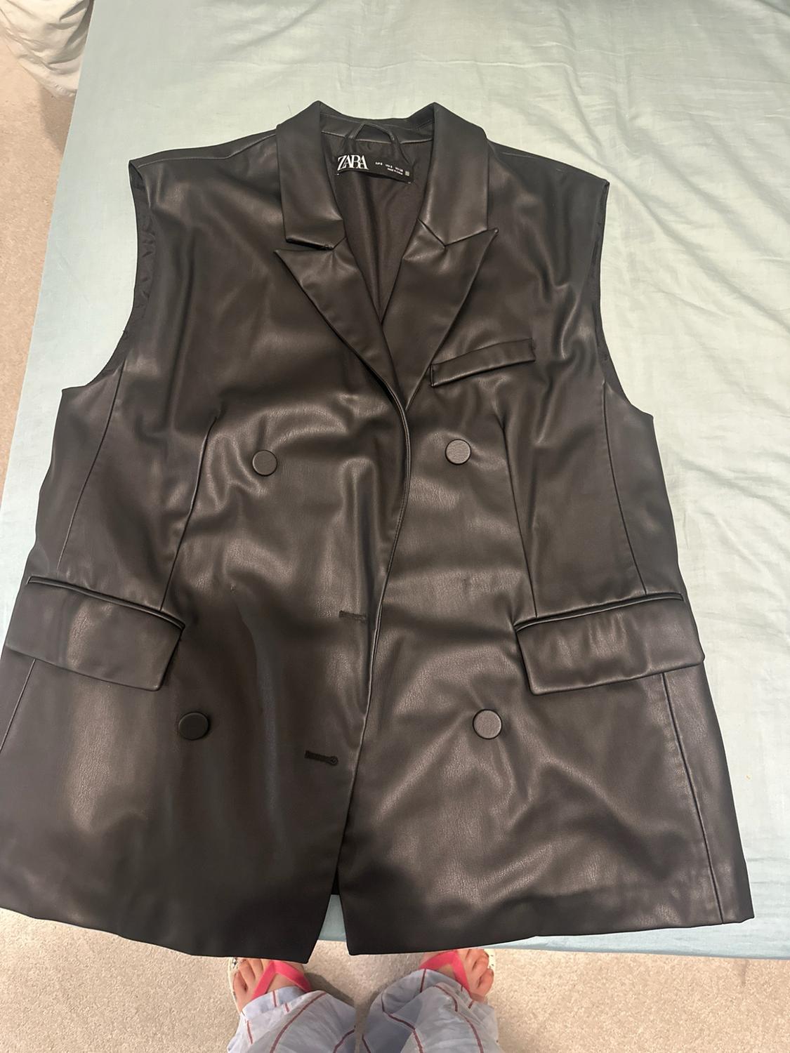 Black waistcoat with a lapel collar, buttons and pockets, on a light blue sheet. Feet can be seen below. Text ZARA.