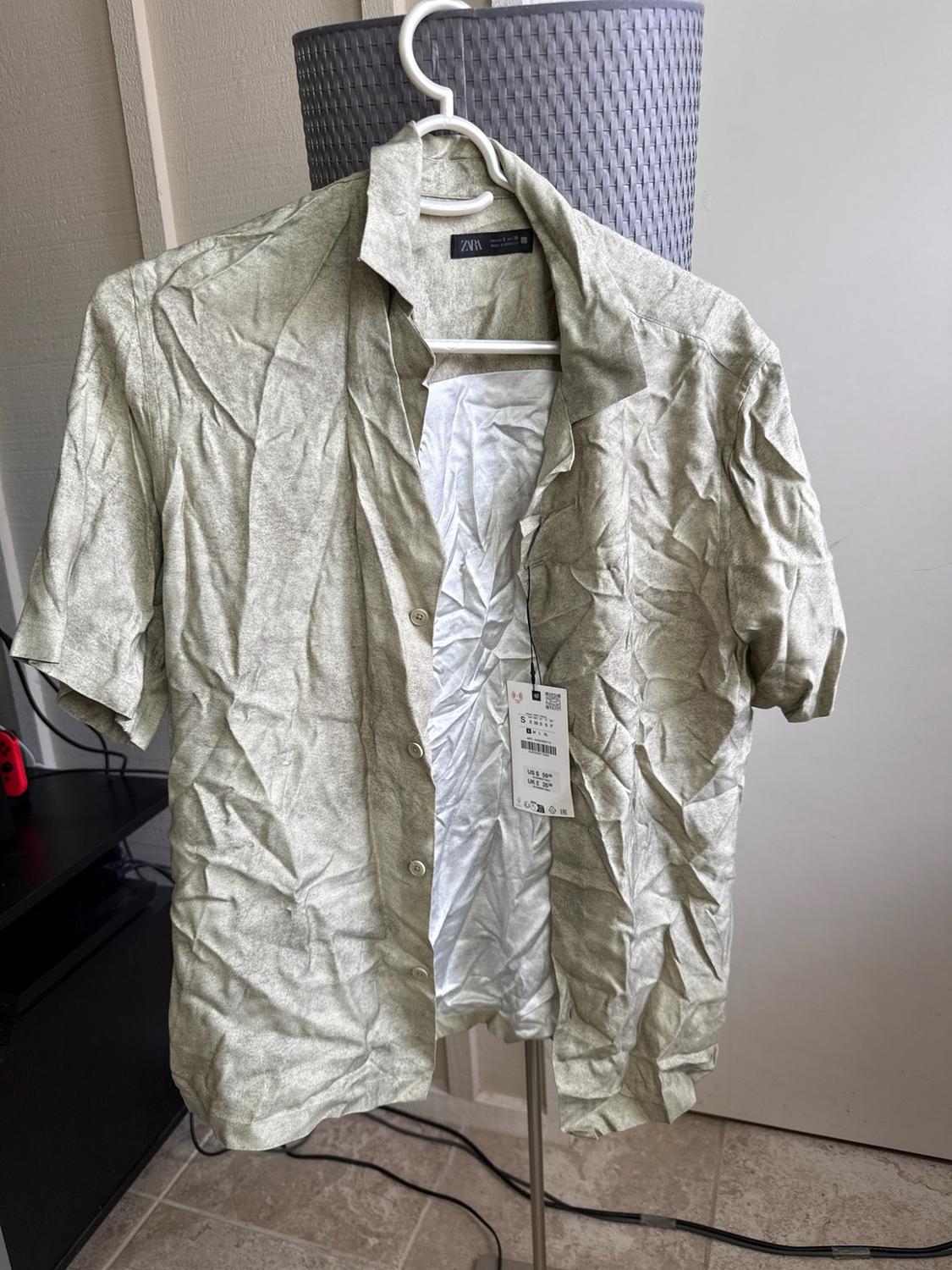 Wrinkled short-sleeved shirt in a light color on a white hanger. It is unbuttoned, showing the ZARA label and a price tag.