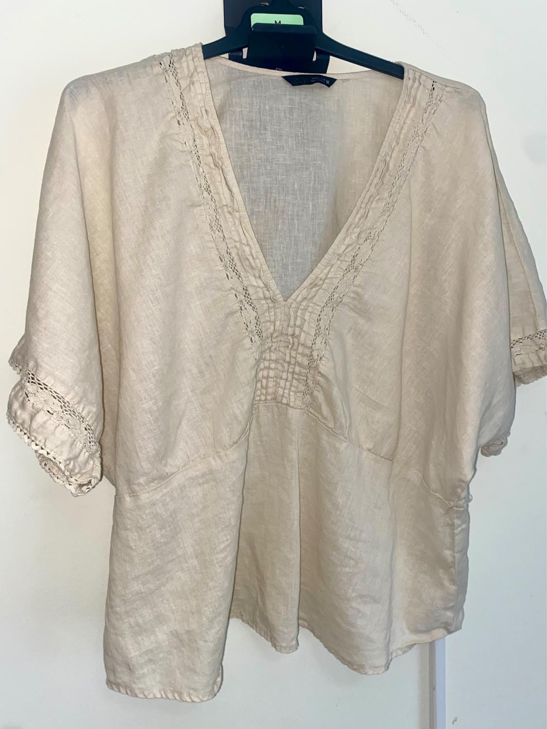 Beige, short-sleeved blouse with a V-neck and lace detailing on the trim, hanging on a black hanger.
