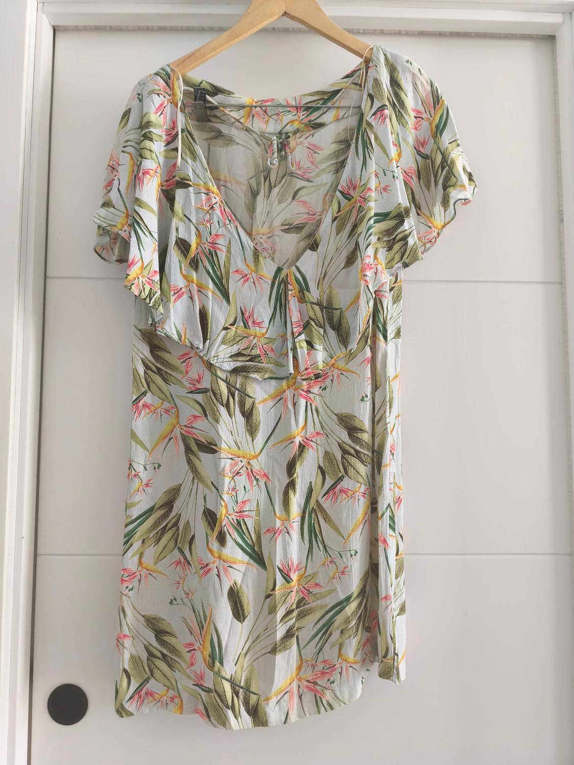 Dress hanging on a hanger, with a tropical floral print in shades of green, pink, and yellow, on a white background.
