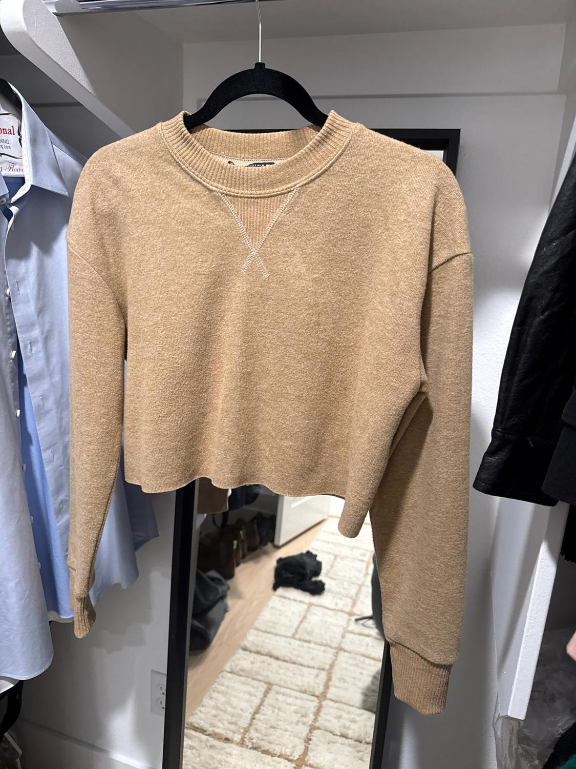 Beige knit short-sleeve sweater with a crew neck, V-shaped detail, and a hem with no finishing, hanging in a closet in front of a mirror.