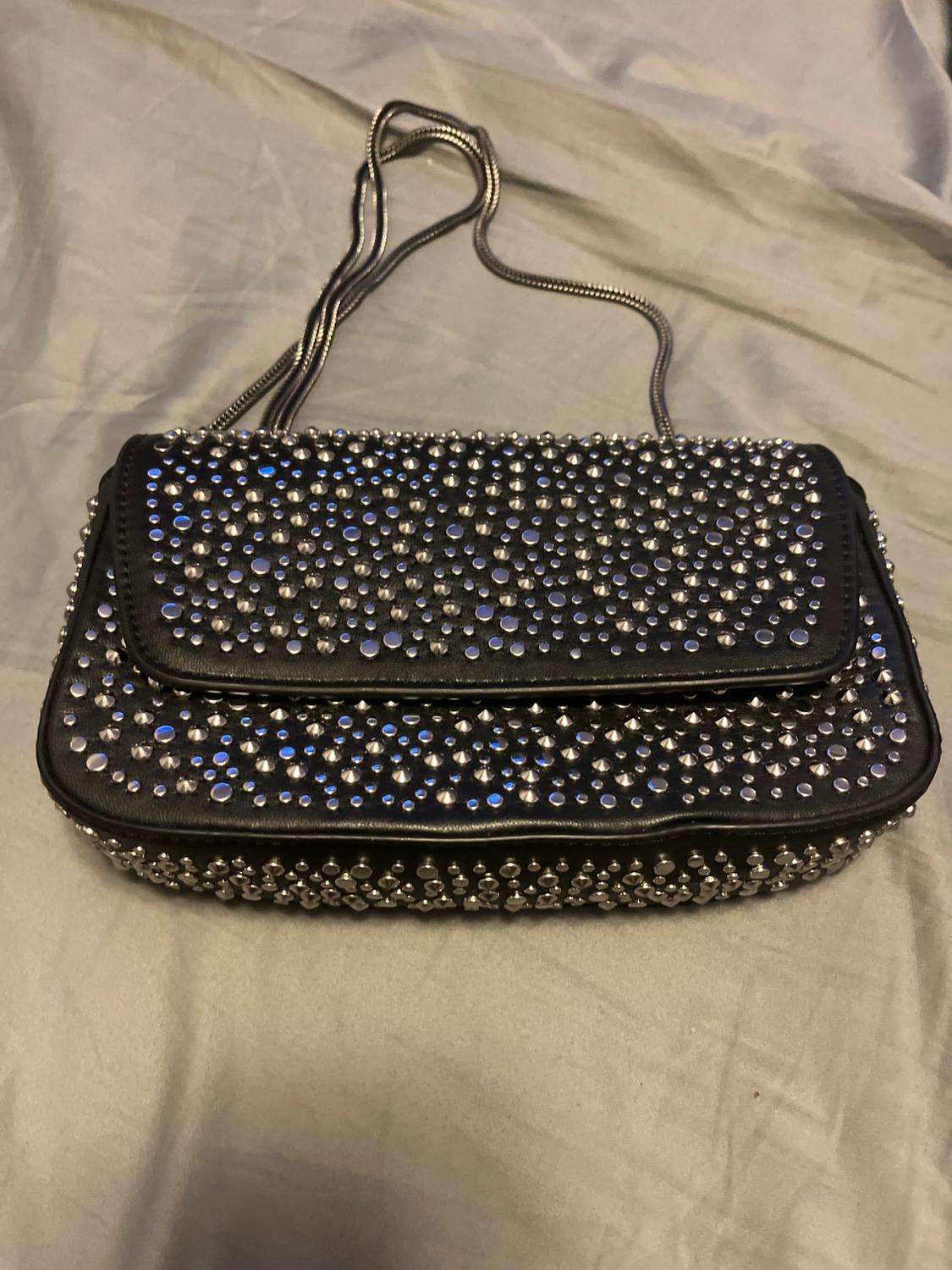 Black handbag with silver studs and metal chain on grey fabric. Stylish and modern design.