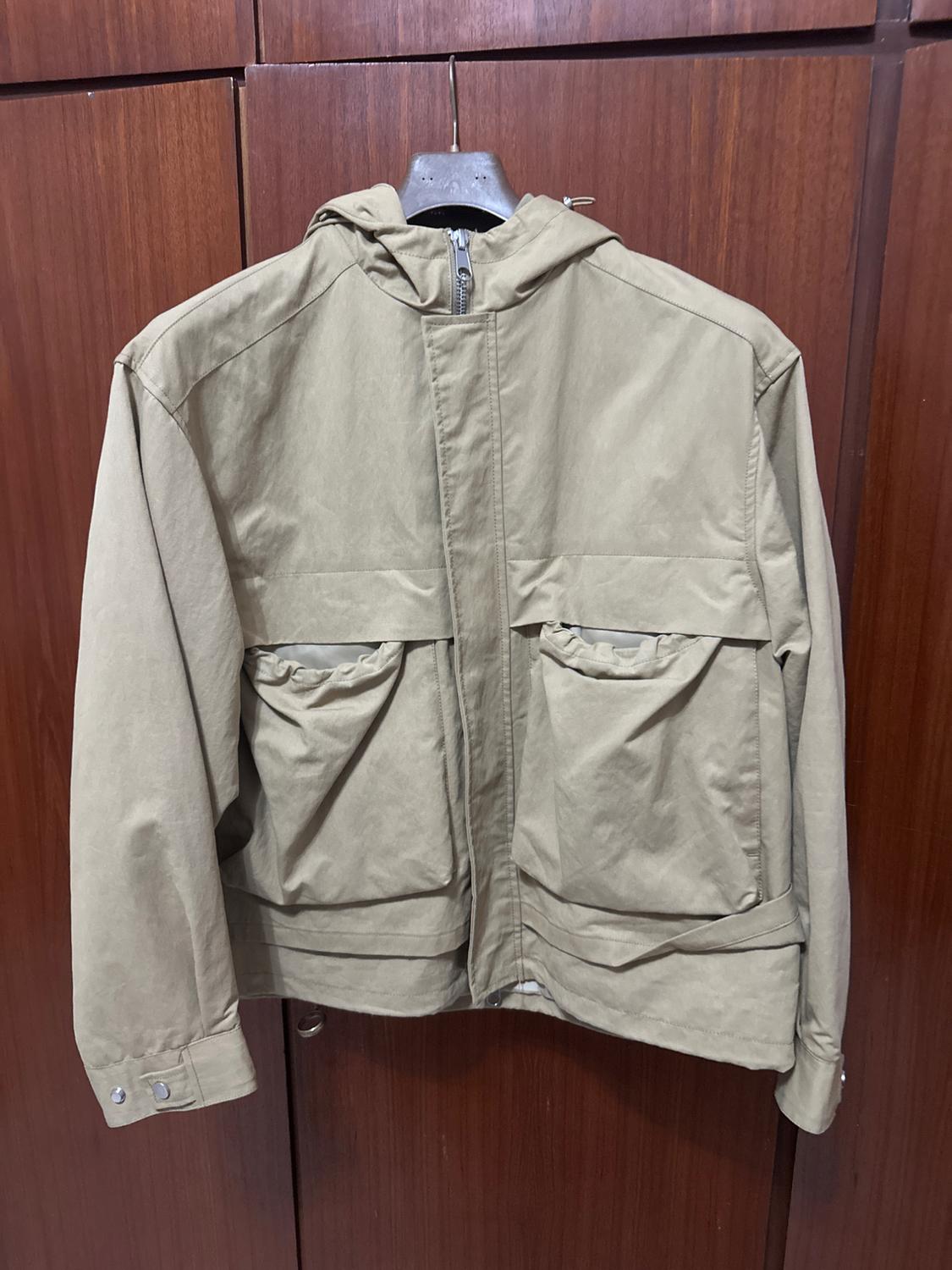 Beige jacket with a hood and large pockets, hanging on a hanger in front of a brown wooden background.