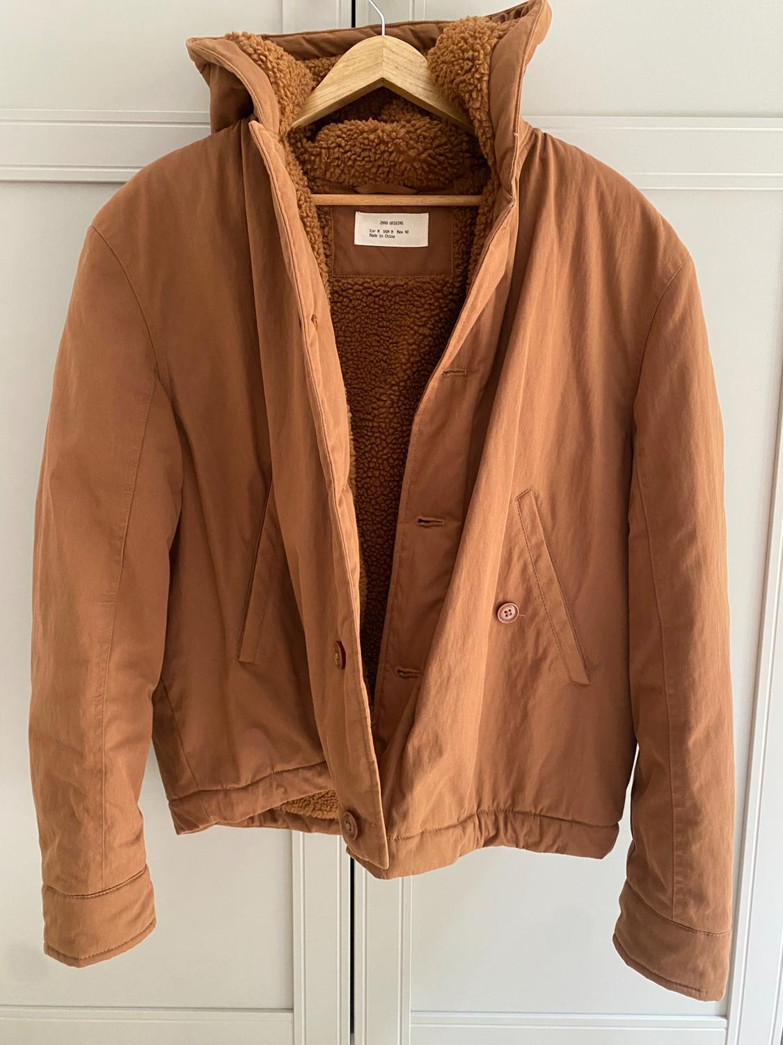 Brown jacket with a wool interior, hanging on a wooden hanger. The label can be seen on the inside of the collar.