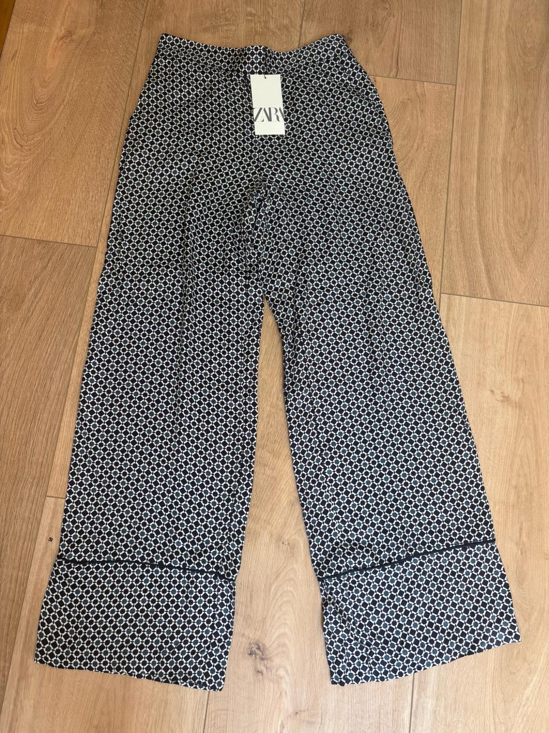 Wide-leg trousers with a geometric print laid out on a wooden floor. Label with the text ZARA.