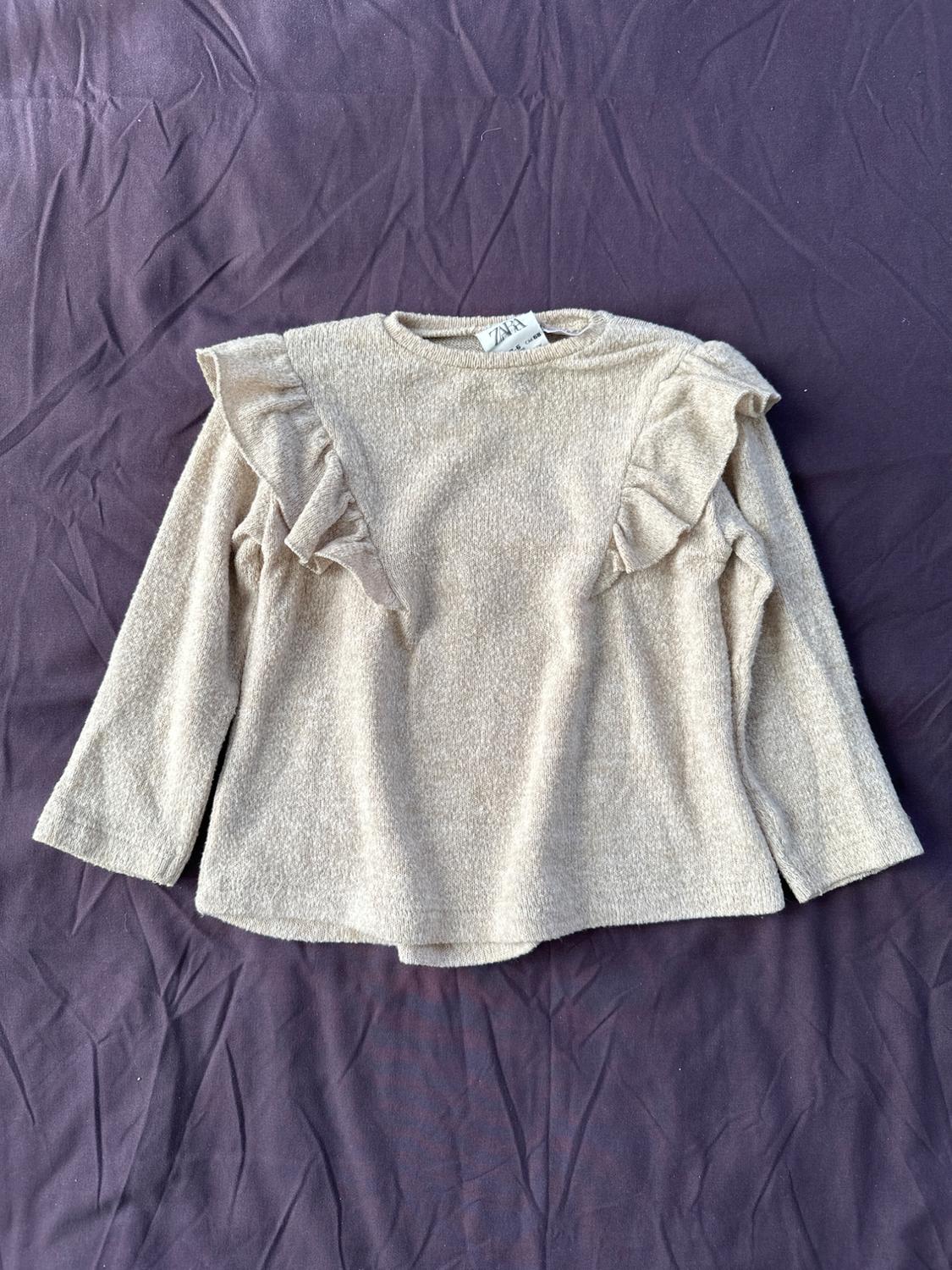 Front view of a beige blouse with ruffles on the shoulders and long sleeves.