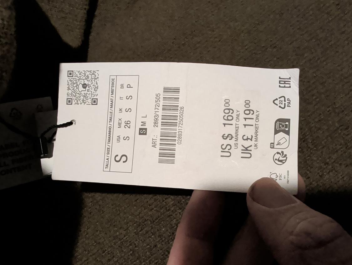 A hand holds a clothing tag showing a price of $169 US and £119 UK, size S, along with a barcode and recycling symbols, over brown fabric.