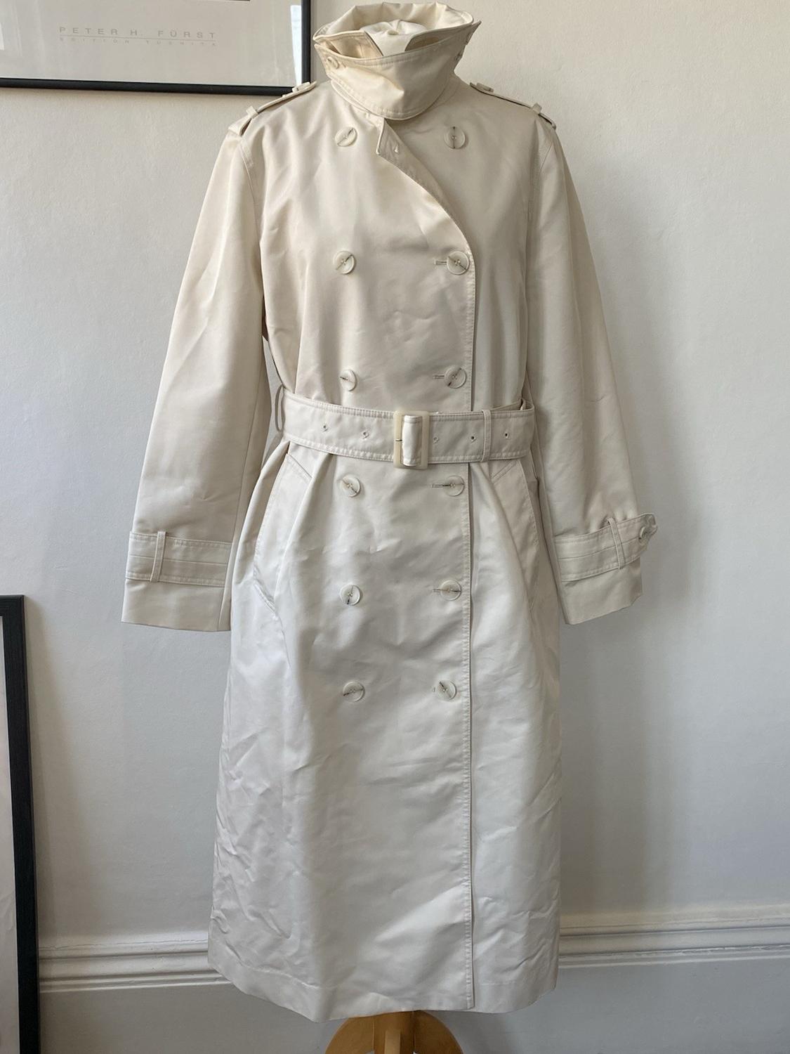 Cream-coloured double-breasted trench coat with belt on a mannequin in a minimalist interior. Text: PETER H. FURST EDITION TUSHITA.