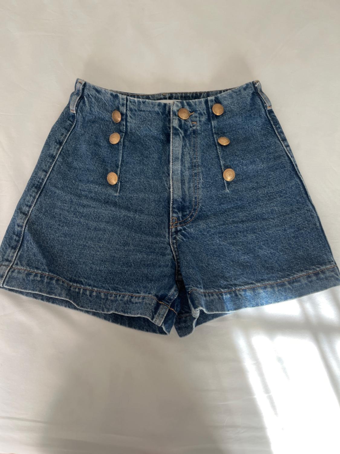 Blue denim shorts with six gold buttons on the front, on a white surface lit by natural light.