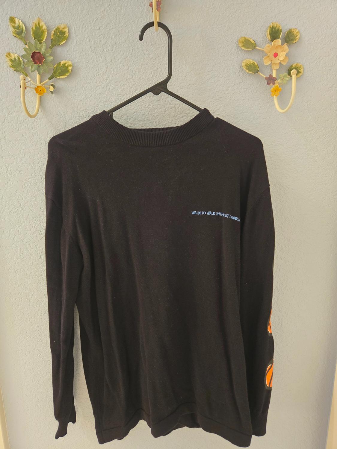 Black long-sleeved jumper hanging on a black hanger. Text on the chest: "WALK TO WALK WITHOUT DRESSING". Floral decoration on the wall.
