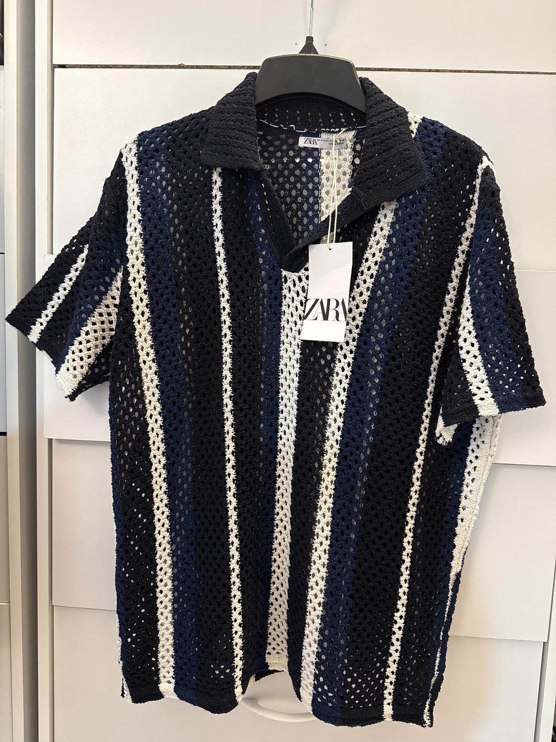 Crochet shirt with vertical stripes in blue, black, and white, hanging on a black hanger. Visible label with the text "ZARA".