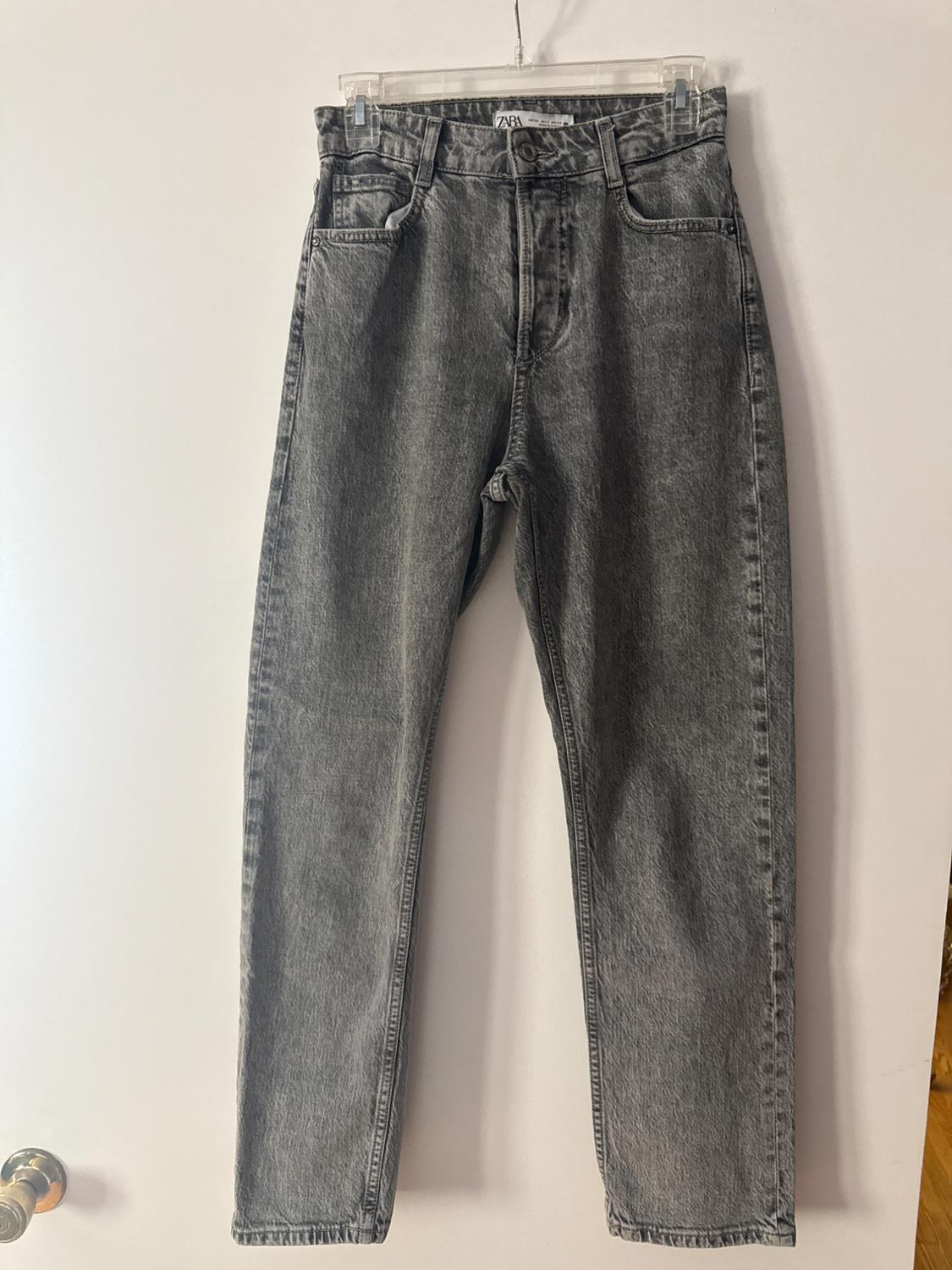 Gray denim pants hanging on a clear hanger over a white door with a silver knob. Inside tag with the text ZARA.