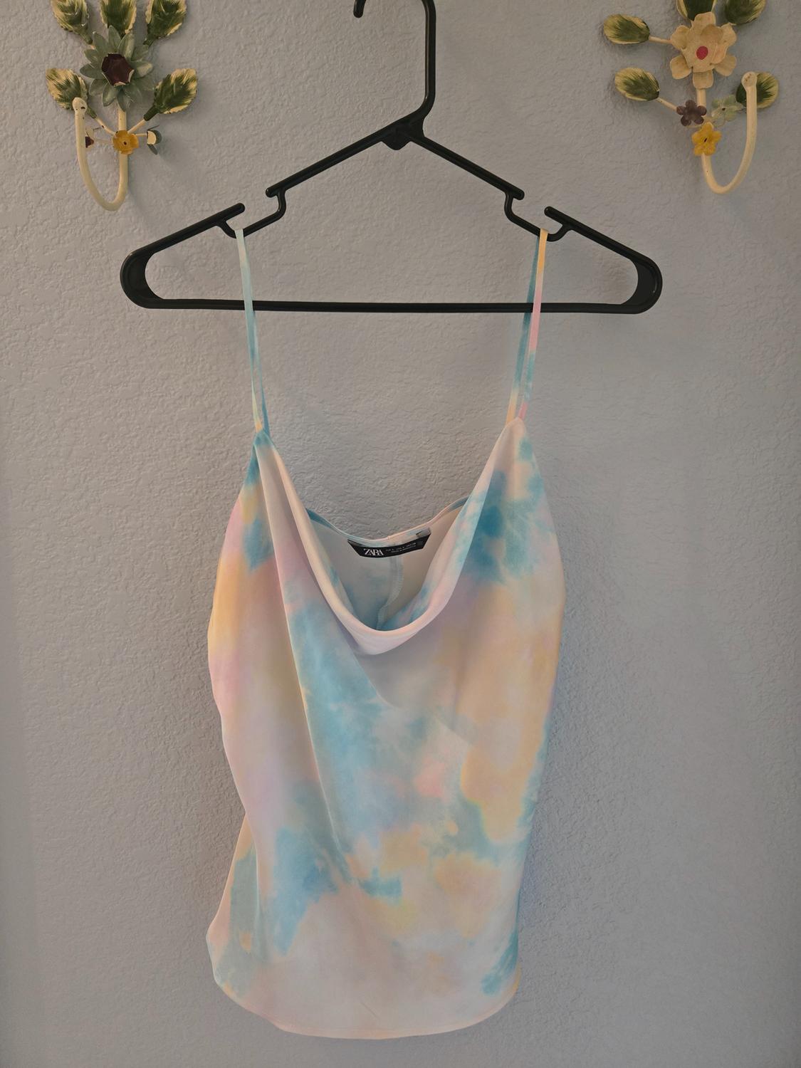 Tie-dye print vest top in pastel shades hanging on a black hanger against a grey wall decorated with flowers.