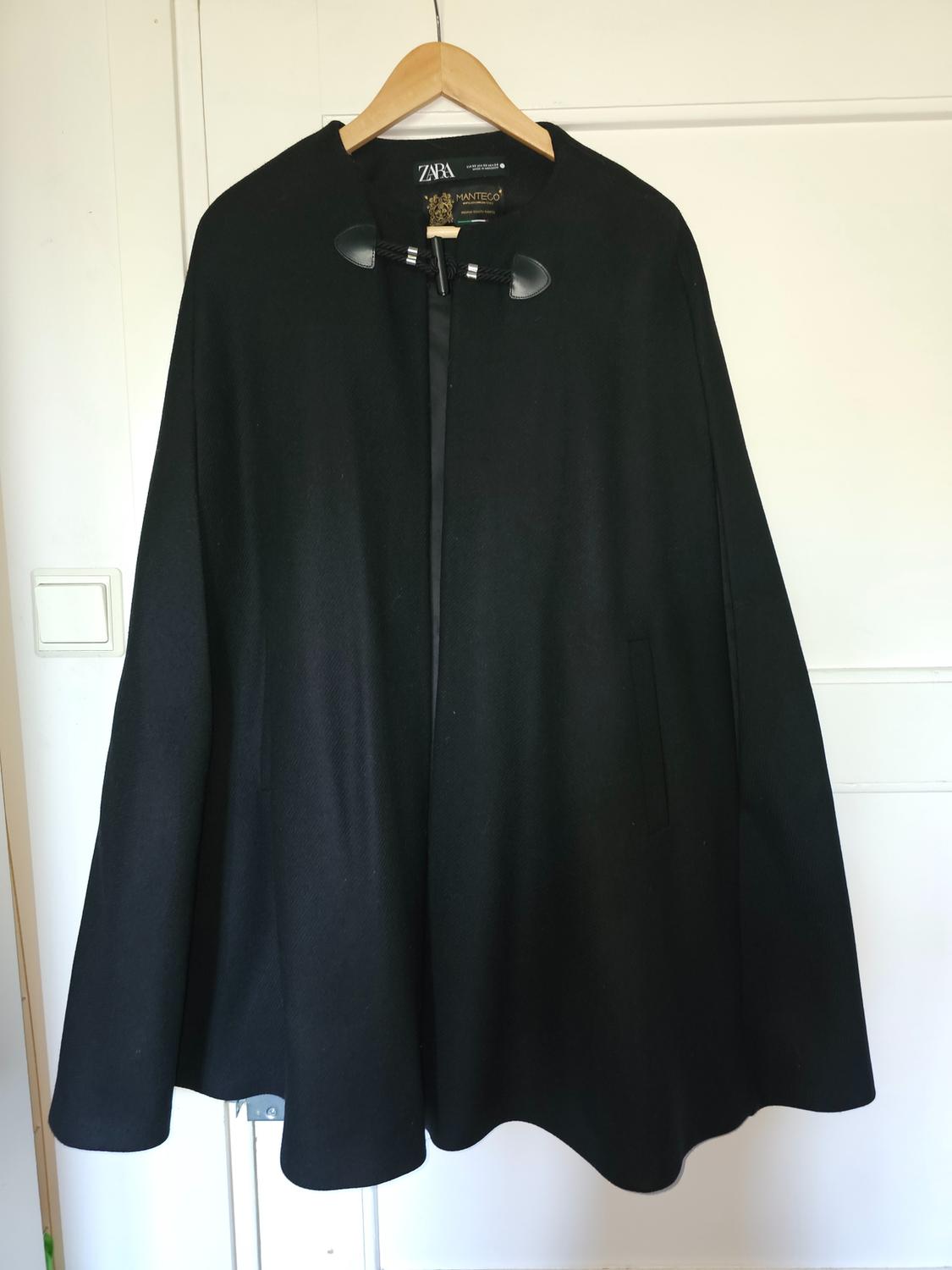 Black cape hanging on a wooden hanger in front of a white door. It has a neck fastening and labels bearing the text ZARA and MANTECO.
