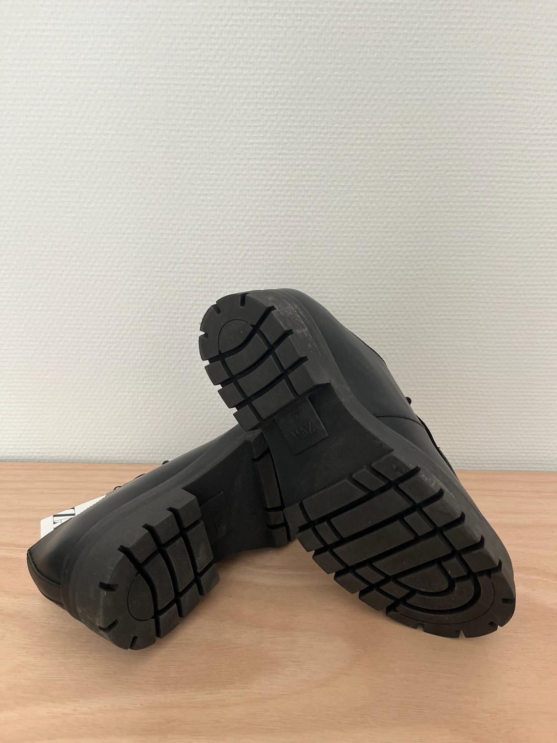 Black shoes with thick, cleated soles on a light wood surface in front of a white wall. ZARA is written on the sole.