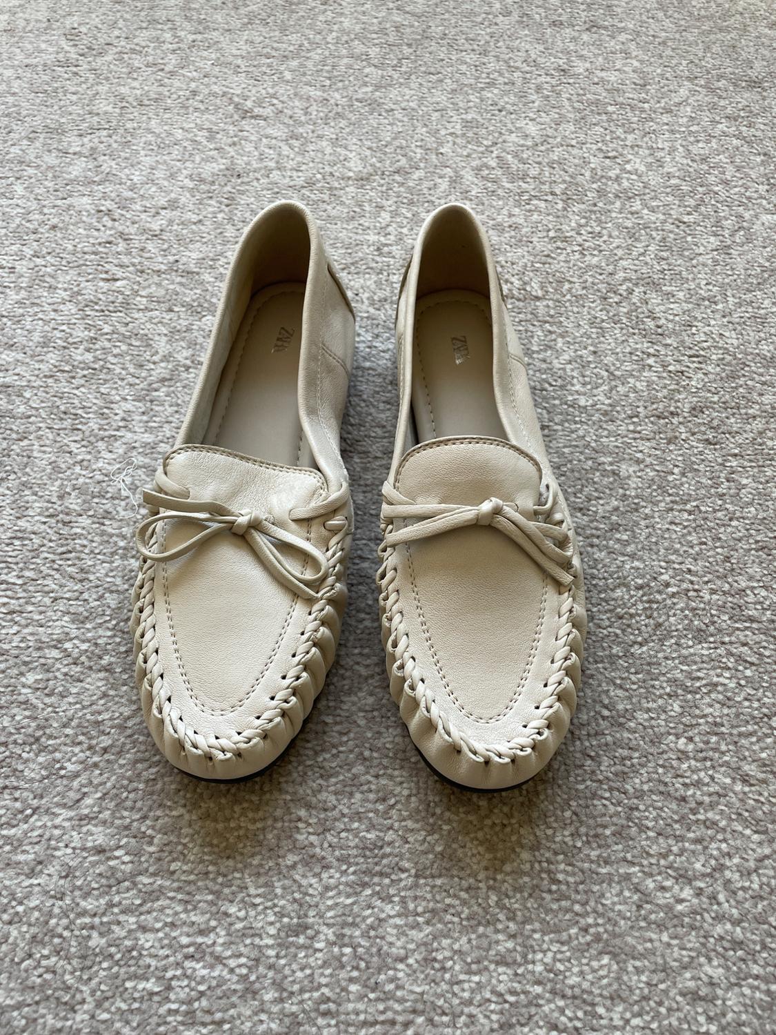 Beige loafers with a bow and braided trim on a light-coloured rug, in a relaxed style. The text 'ZARA' can be seen on the inside.