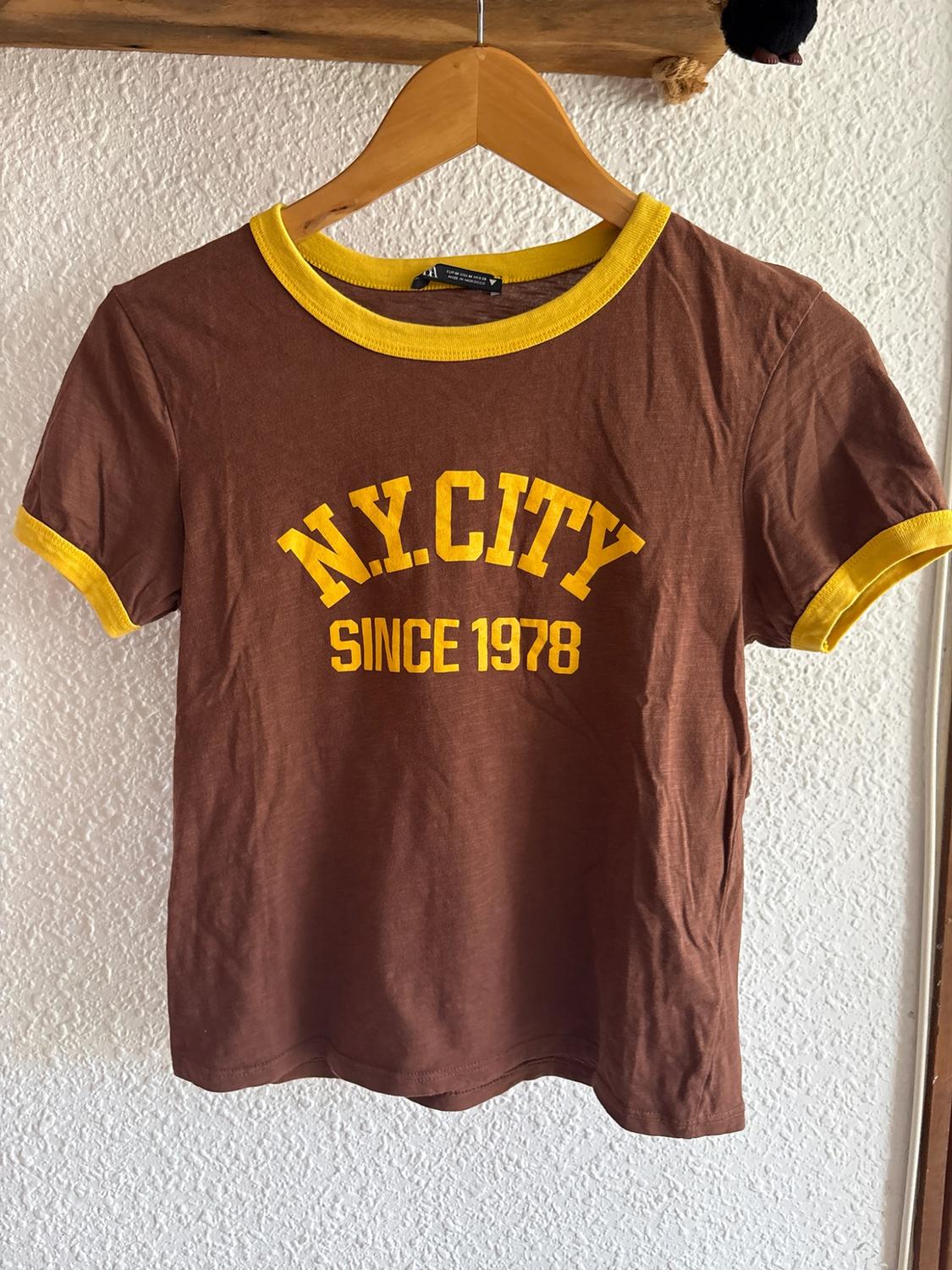 Brown short-sleeved t-shirt with yellow trim on a wooden hanger. Printed text: "N.Y. CITY SINCE 1978". White wall background.