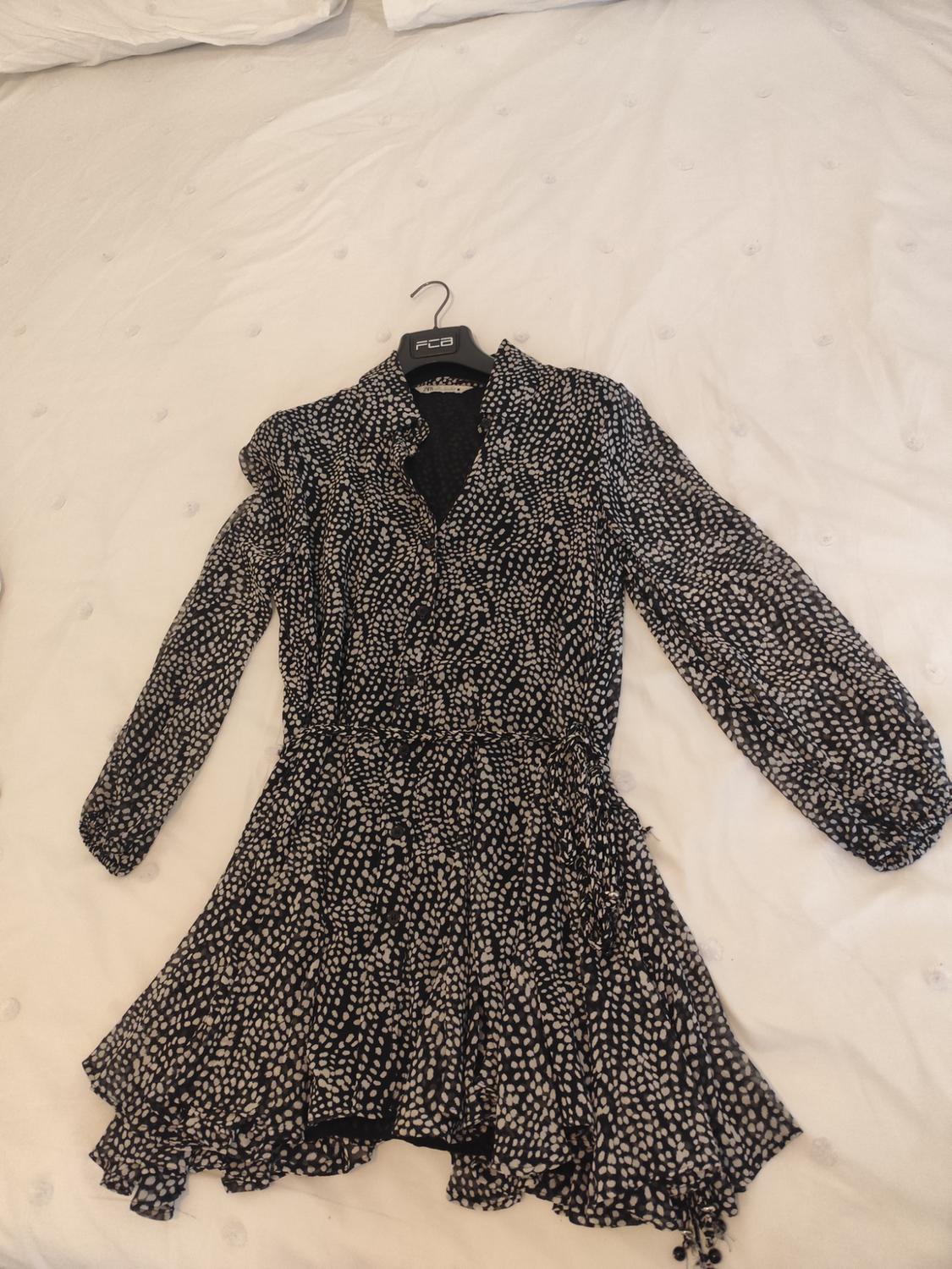 Black dress with white polka dot print, long sleeves and a fitted waist, on a white bed. Hanger with text "FCO".