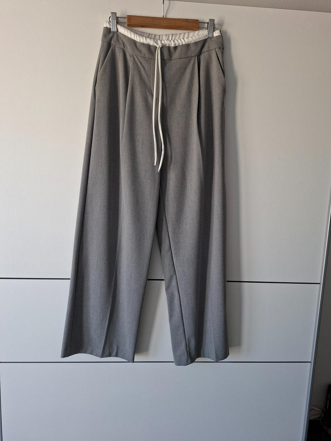 Wide grey trousers with a pronounced crease, hanging on a hanger. Their white elasticated waistband with drawstring stands out. White panelled background.