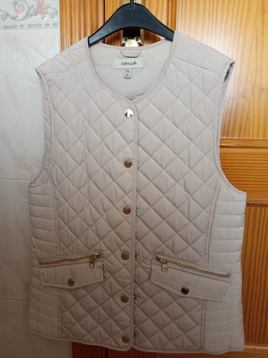 Beige padded gilet with gold buttons and zip pockets, hanging on a black hanger. "ZARA Girls" label visible.