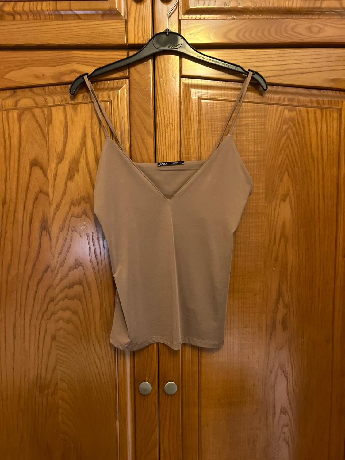 Beige camisole top with double spaghetti straps and V-neckline featuring a ZARA label, hanging on a black hanger in a wooden wardrobe.