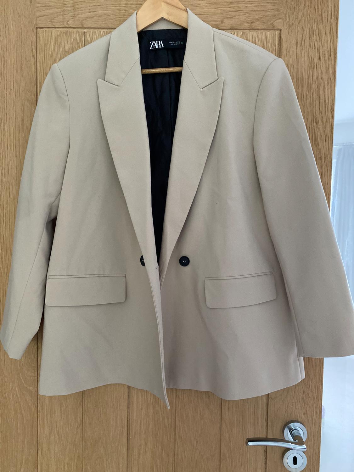 Beige Zara jacket hanging on a wooden door. It has wide lapels and two visible black buttons.
