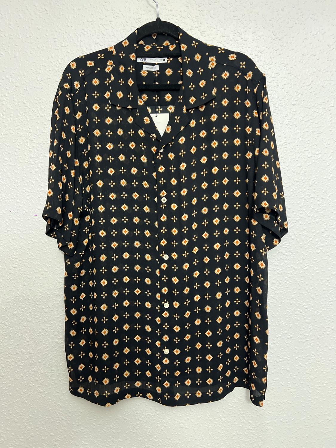 Black short-sleeved shirt with a geometric print in orange and white tones, hanging on a hanger against a white background.
