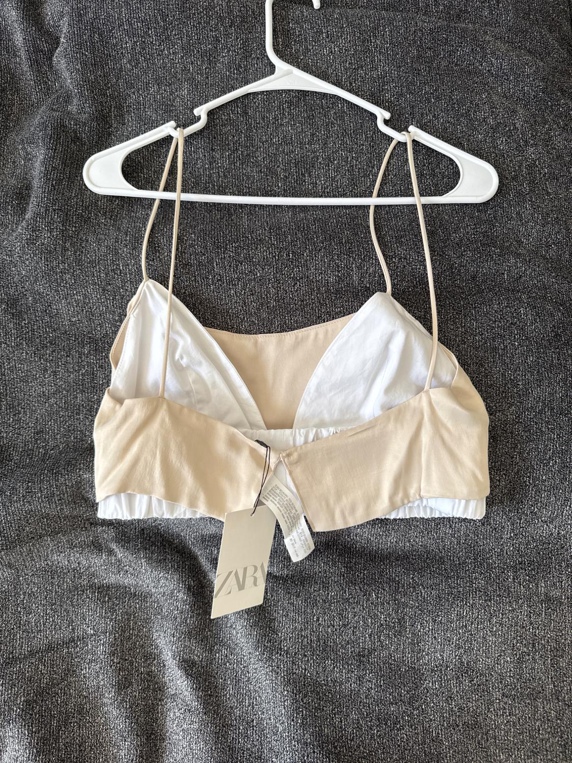 Beige and white bra hanging on a white hanger over gray fabric. Visible label with the text "ZARA".