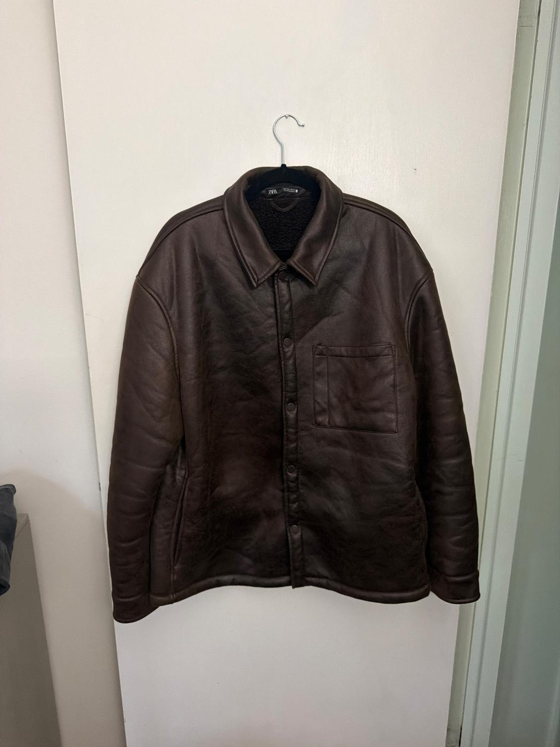 Dark brown jacket hanging on a hanger on a white door. Design with a collar, button front closure, and a chest pocket.