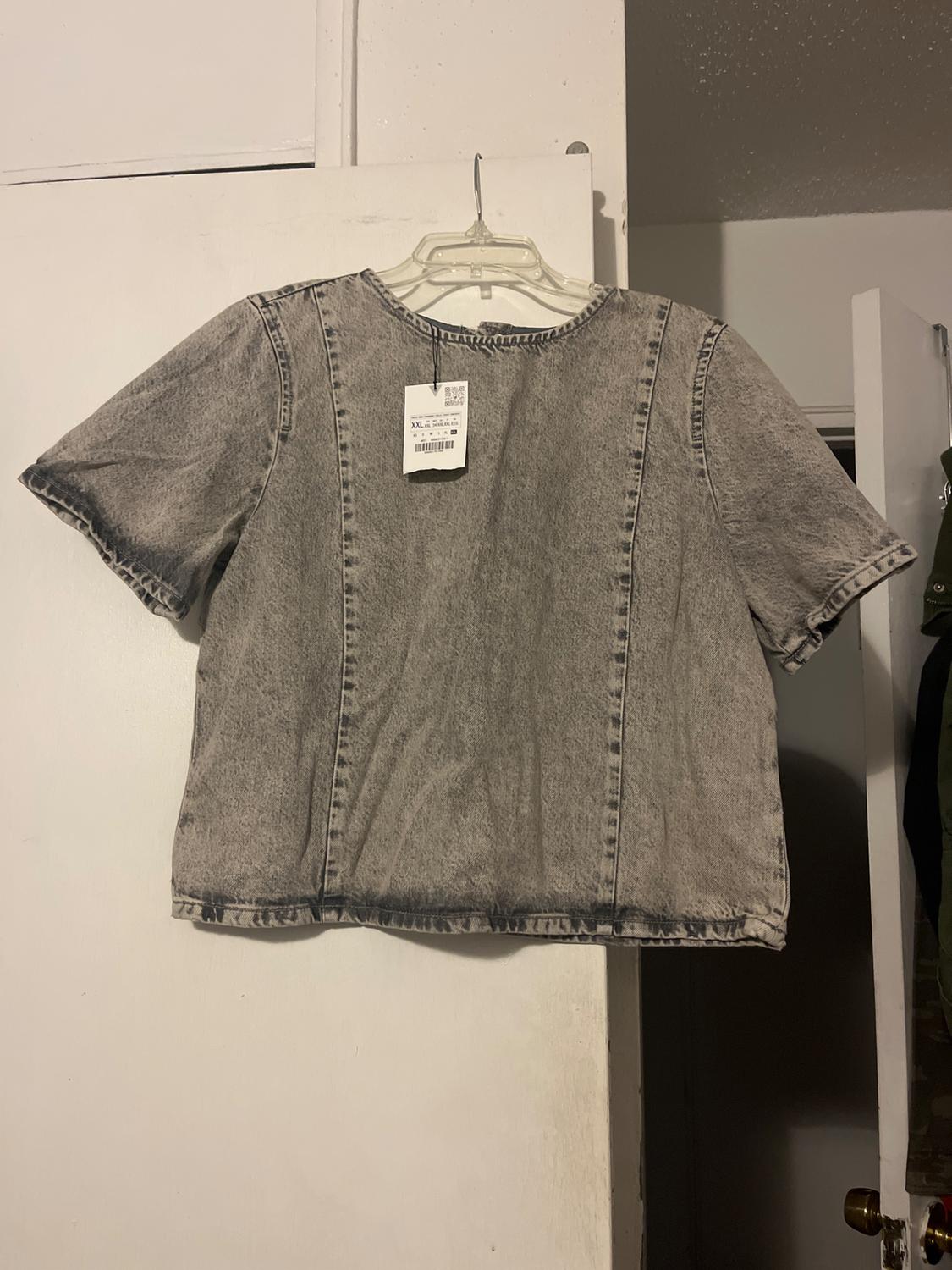 Grey denim T-shirt hanging on a transparent hanger, with visible label, on a white door in an indoor environment.