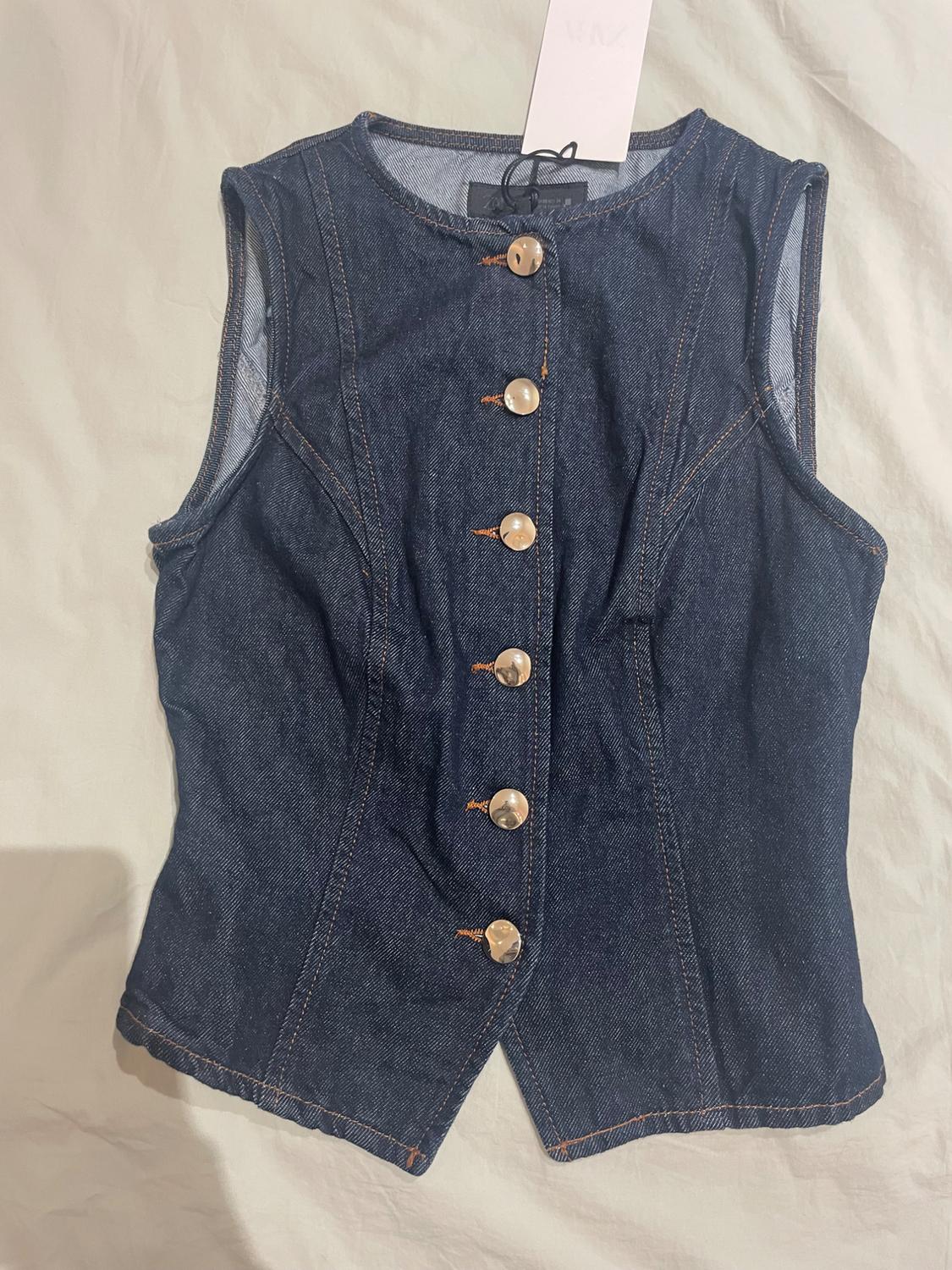 Sleeveless blue denim waistcoat with gold front buttons on a light background. White tag hanging from the collar.