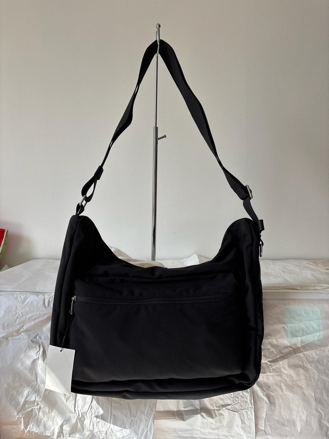 Black bag with a front pocket hanging from a metal stand on crumpled white paper. Includes a label with the text ZARA.