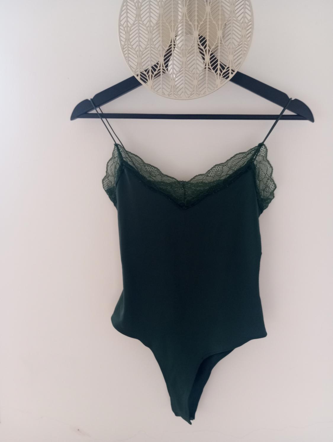 Dark green bodysuit with lace, hanging on a black hanger, white background with embossed leaf decoration.