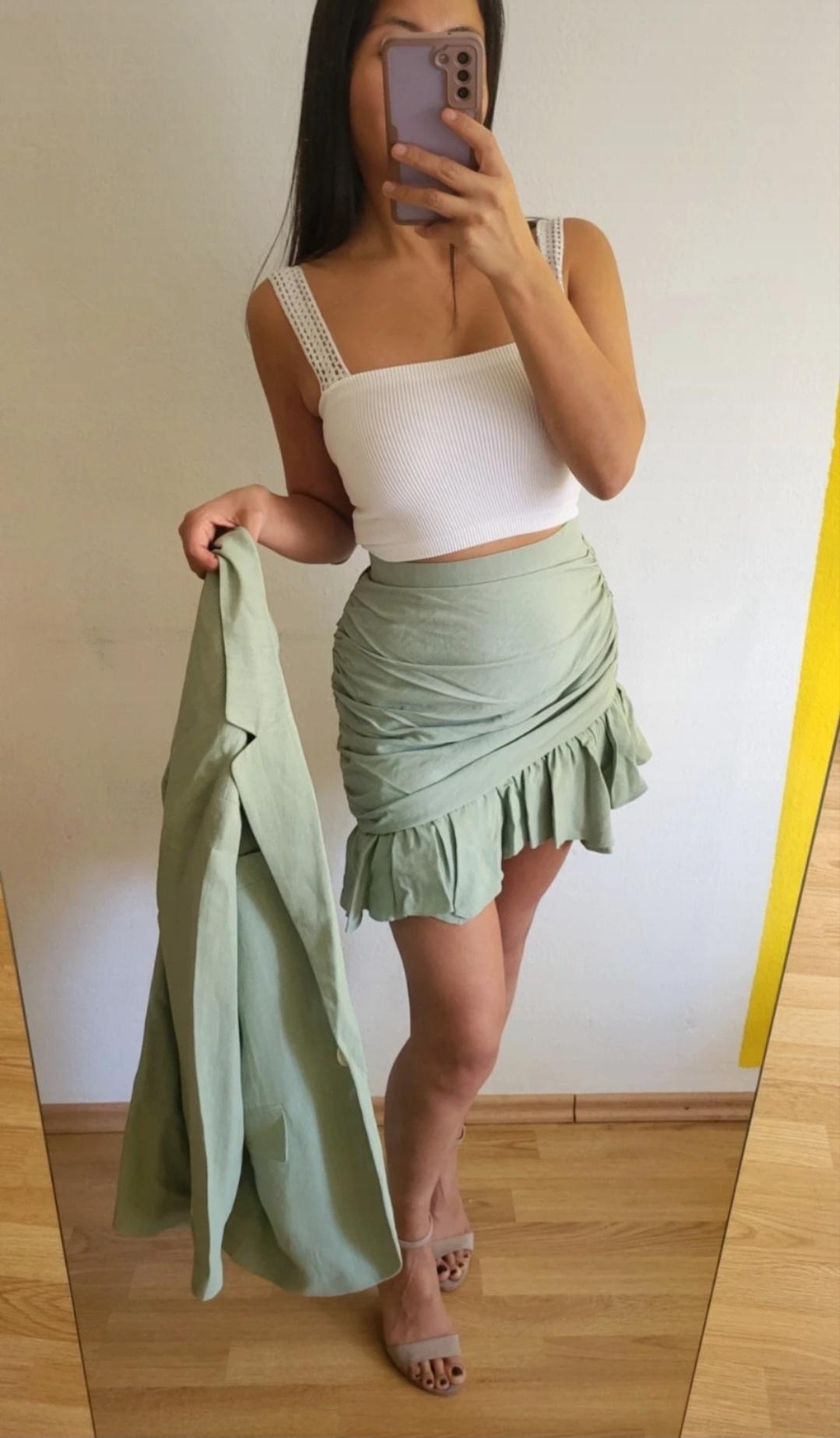 Woman in front of a mirror wearing a white vest top, green skirt with frills, green blazer in hand and beige sandals. White and yellow background.