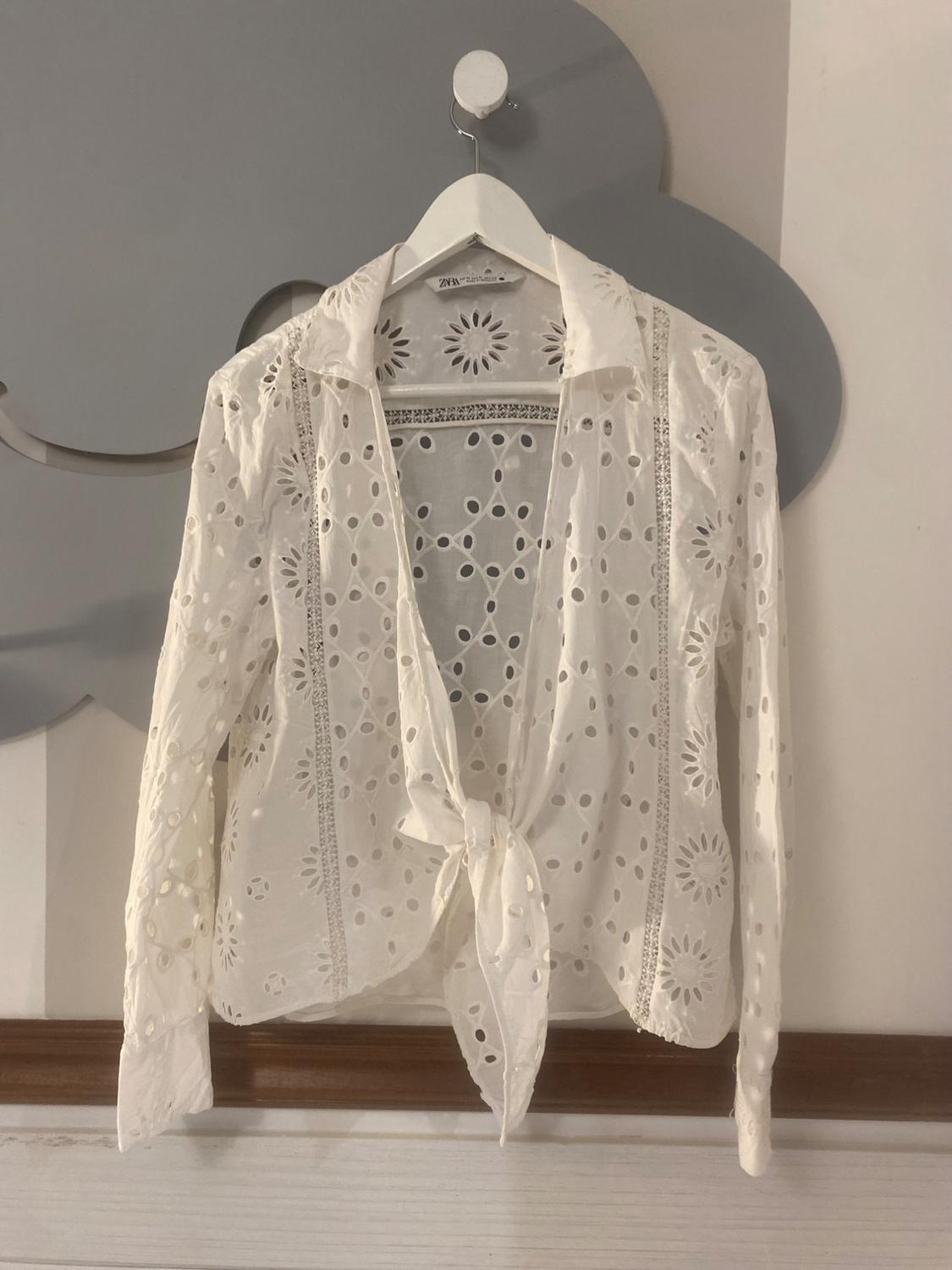 White long-sleeved blouse with openwork embroidery and front knot, hanging on a hanger in front of a grey cloud decoration. ZARA label.