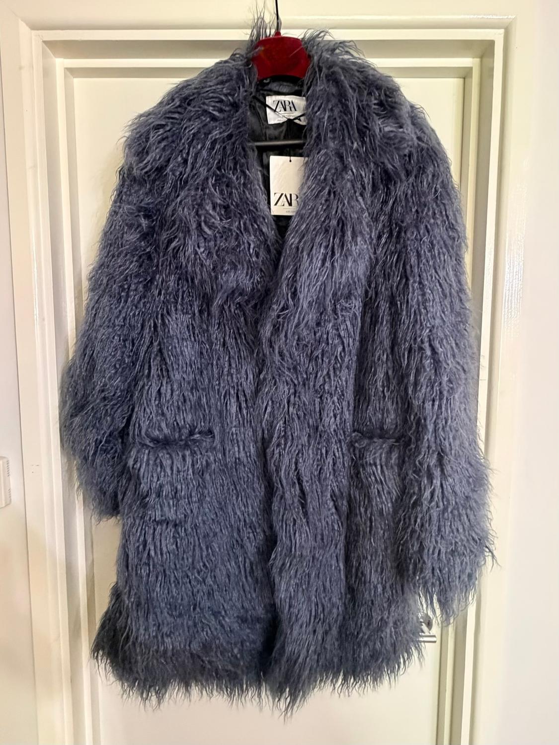 Grey fur coat hanging on a red hanger, with a visible label that reads "ZARA", on a white door.