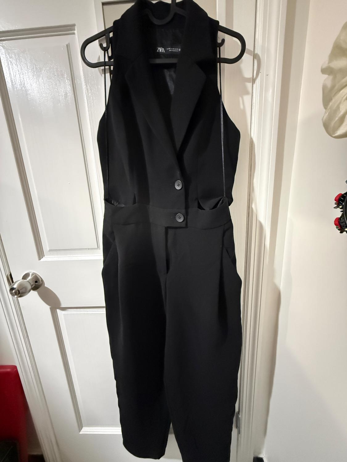 Sleeveless black jumpsuit hanging on a hanger in front of a white door. The label says "ZARA".