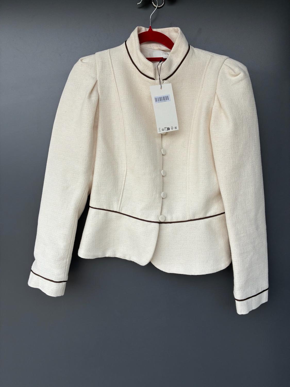 Cream high-neck jacket with dark trims, buttons and puffed shoulders, hanging on a red hanger against a dark grey background.