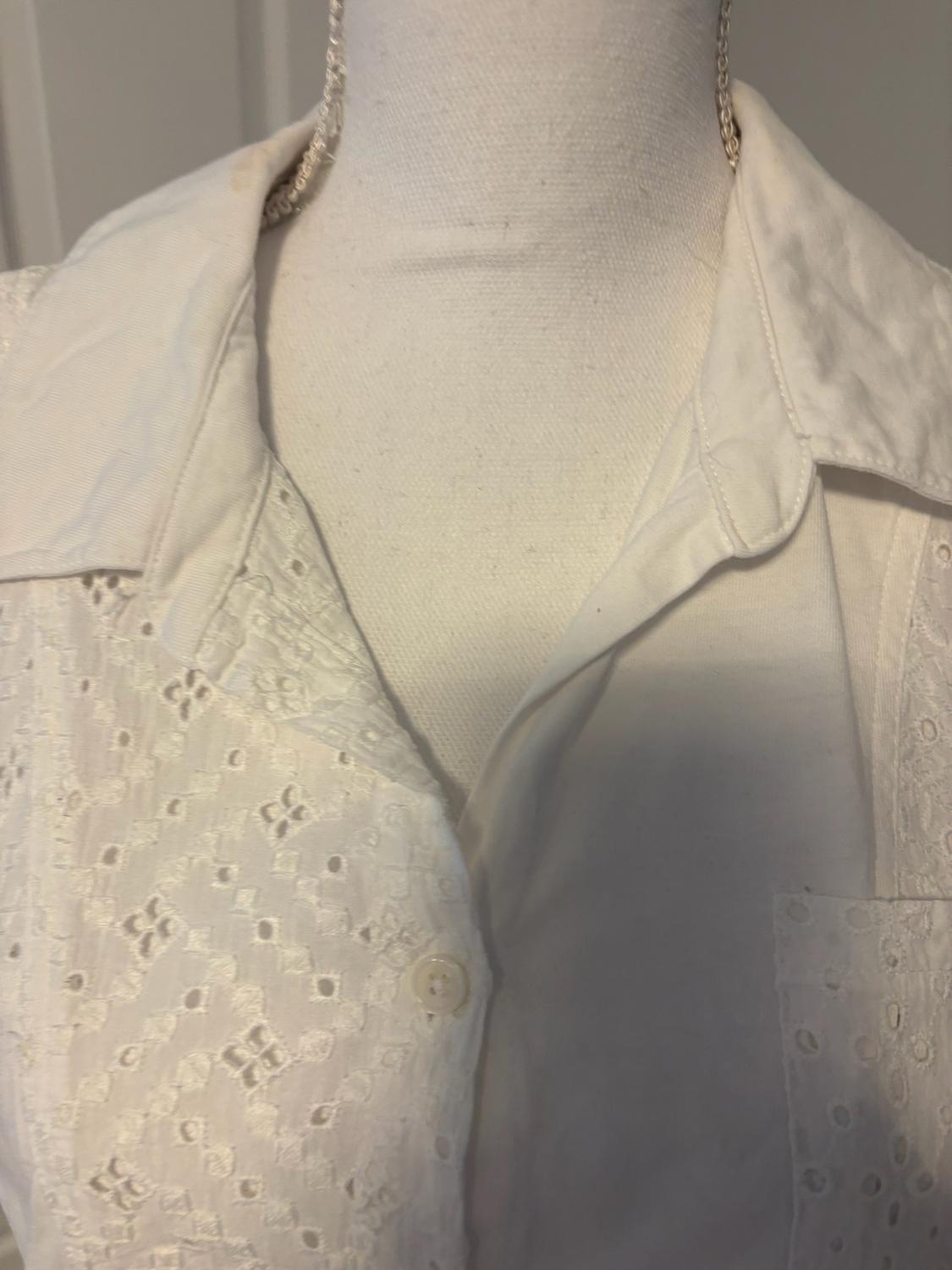 Detail of a white shirt on a mannequin, with an open collar, button, and fabric with broderie anglaise on the front and pocket.