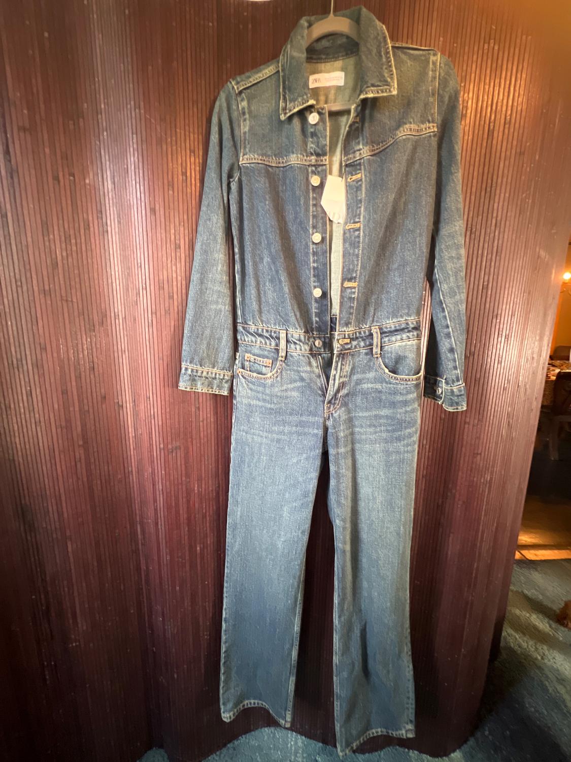 Blue denim jumpsuit hanging on a hanger in front of a brown wooden background.