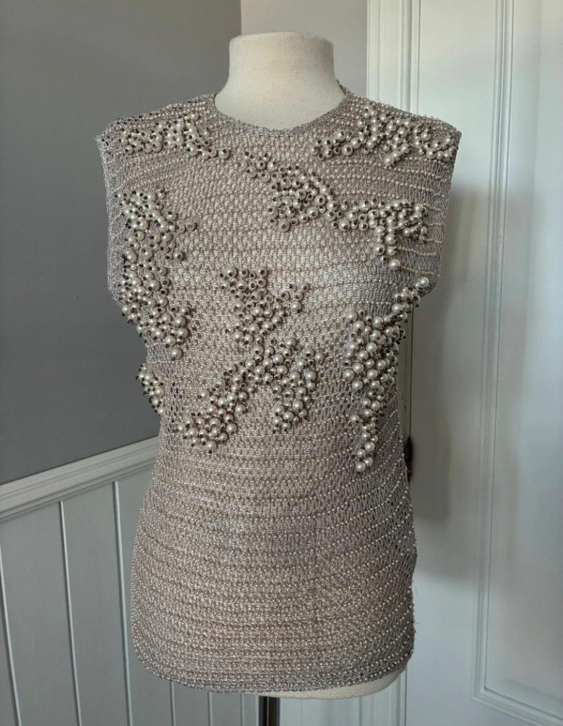 Mannequin with a beige knitted top, decorated with pearls in floral patterns. White and grey wall background.